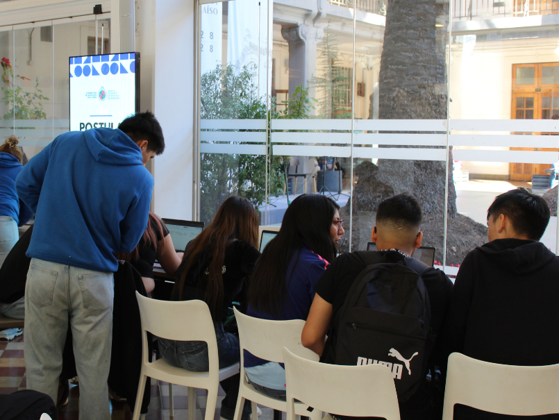Estudiantes postulan a través de computadores, en Casa Central PUCV. Se ven de espaldas, conversando y trabajando en computadores.